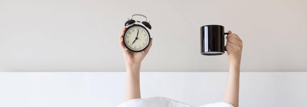 Woman showing arm raised up holding coffee cup and black alarm clock behind duvet in the bed room, Young girl with two hands sticking out from the blanket. wake up with fun in morning concept.
