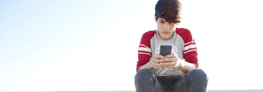 Attractive and thoughtful teenager boy relaxing with a skateboard and sitting down on a bench by the sea, holding and using a smartphone for networking during a sunny day, outdoors.